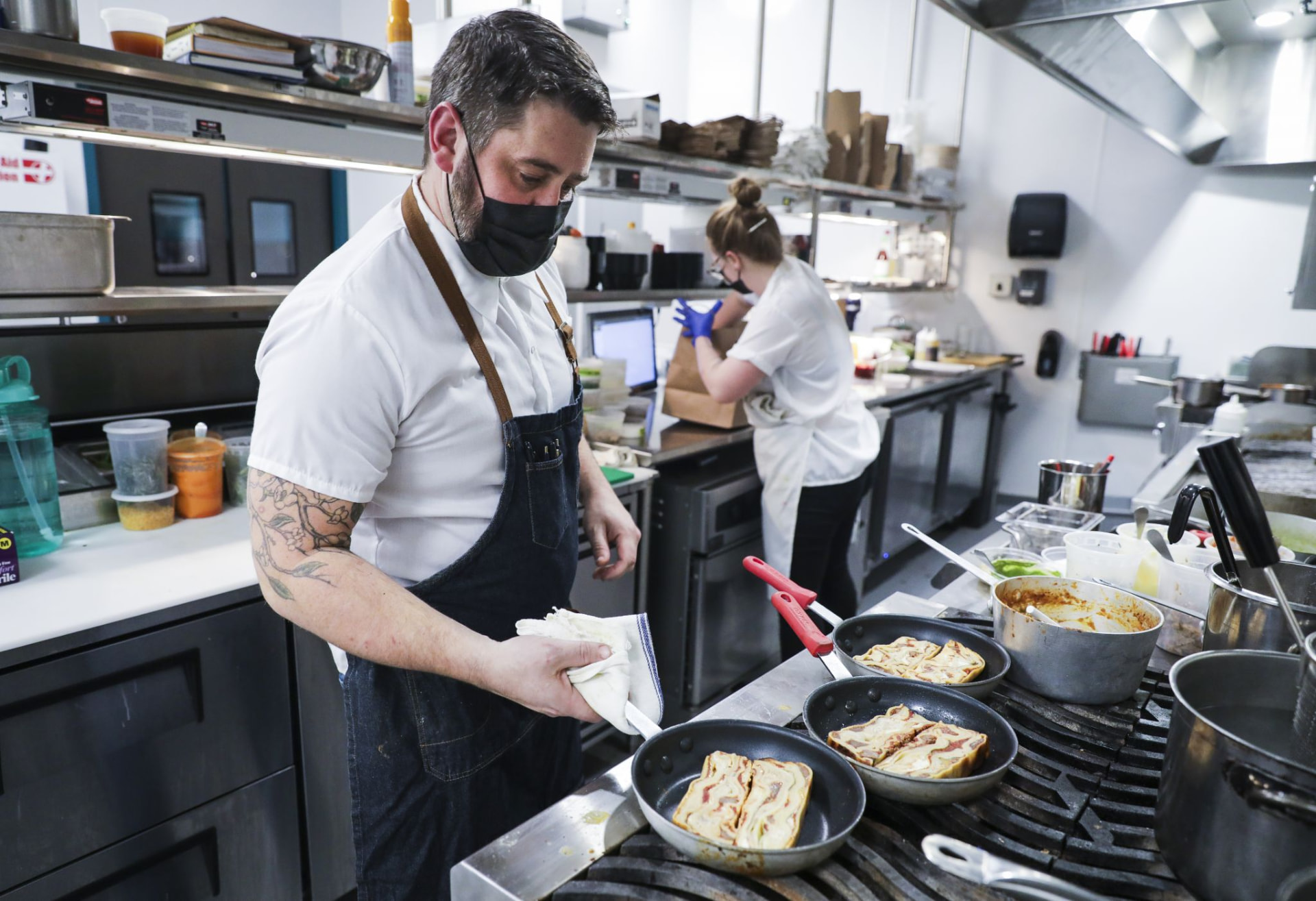 A masked cook working several pans across a hot range during service
