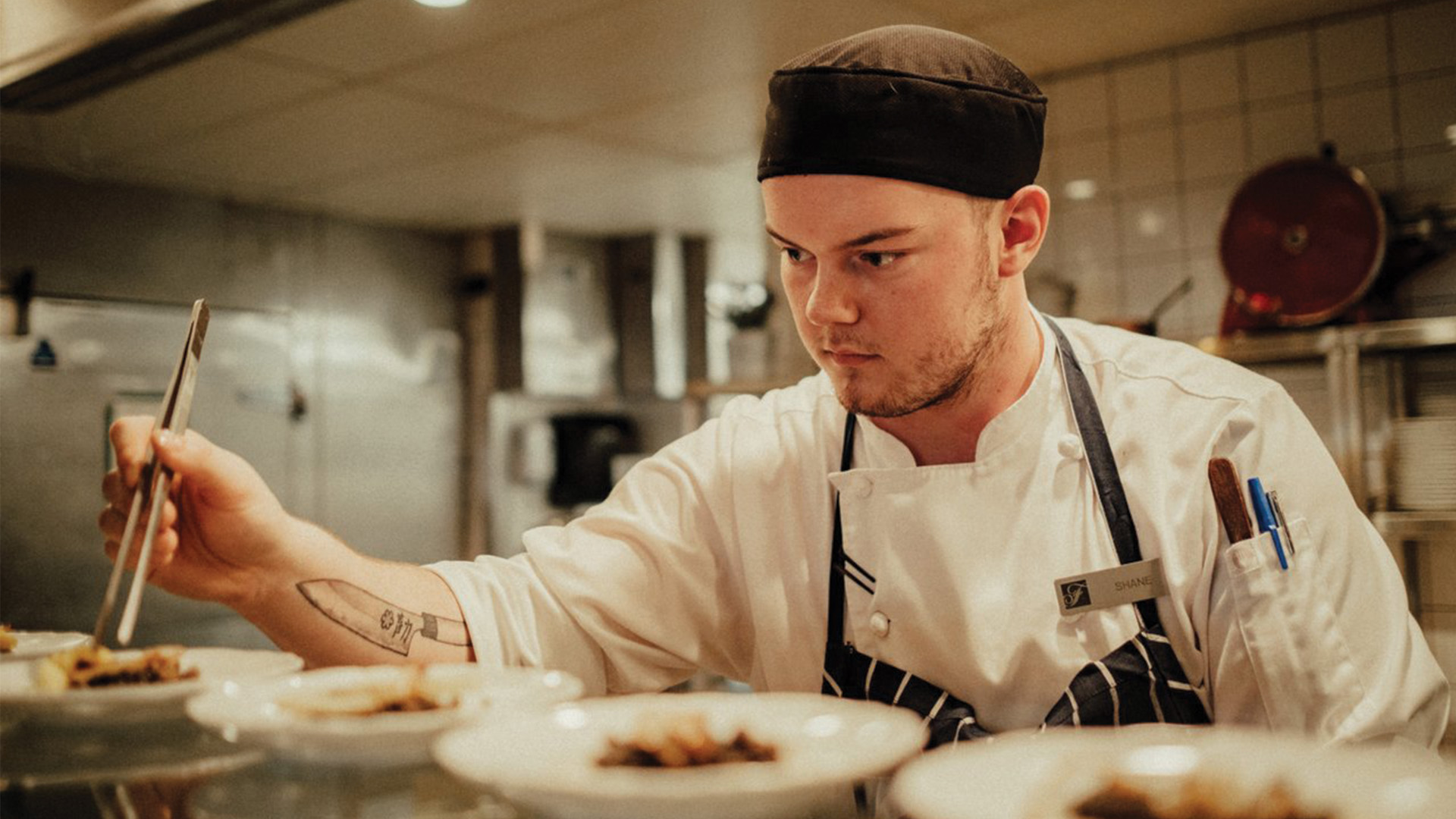 A chef placing a garnish with tweezers, inches from the plate